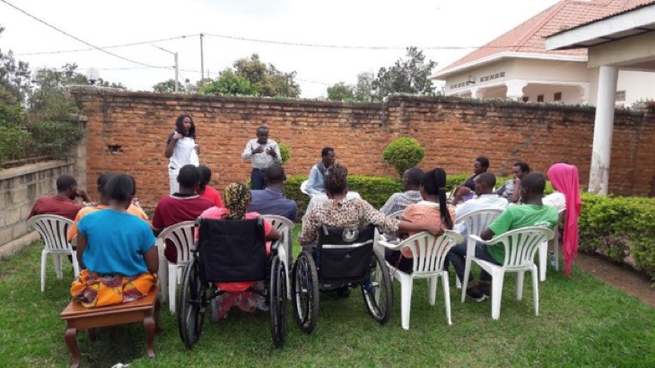 A photo of a group of young people taken from behind. They are sitting on chairs and in wheelchairs while two people who are gesturing stand in front.
